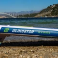 Gladiator Origin 10.6 – logo na burcie deski SUP przy brzegu, ujęcie lifestyle z wodą, mostem i górami w tle