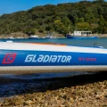 deska sup Gladiator Origin 10.8 Rental – logo Gladiator na burcie, ujęcie lifestyle nad wodą z łodzią w tle