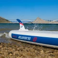 sup Gladiator Origin 10.8 Rental – burta i statecznik deski na kamienistej plaży, ujęcie lifestyle z falą i mostem w tle