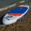 sup Gladiator Origin 10.8 Rental – deska SUP na kamienistej plaży, pad EVA, linki bagażowe i zawór, ujęcie przy fali