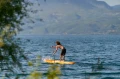 pływanie na desce SUP Aqua Marina Fusion po jeziorze otoczonym górami widziane zza gałęzi drzew